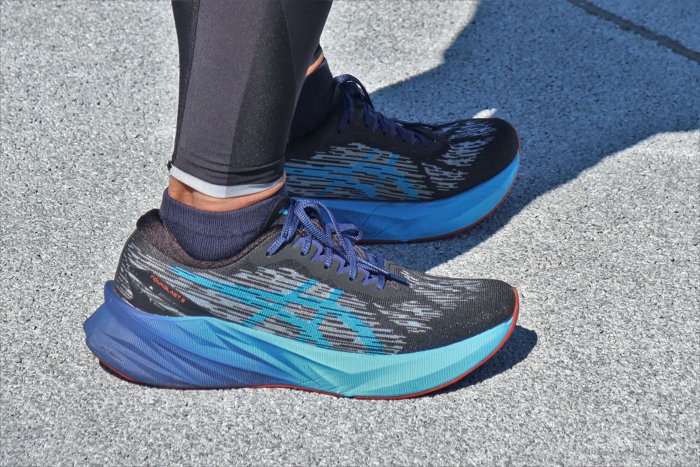 A person's feet are standing on a sidewalk with a pair of blue running shoes.