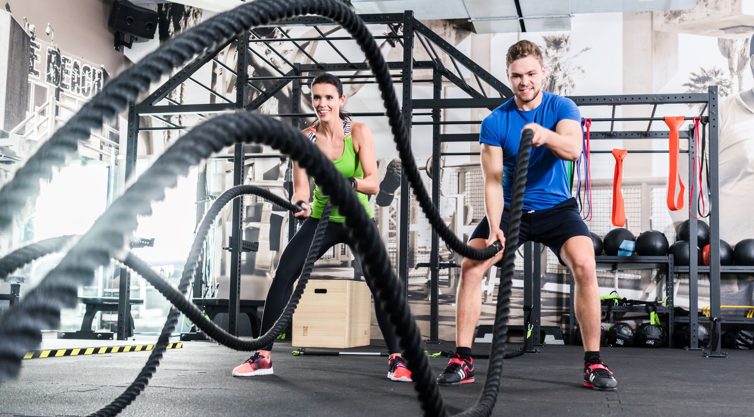 Two people exercising in a gym, one using battle ropes and the other using resistance bands.