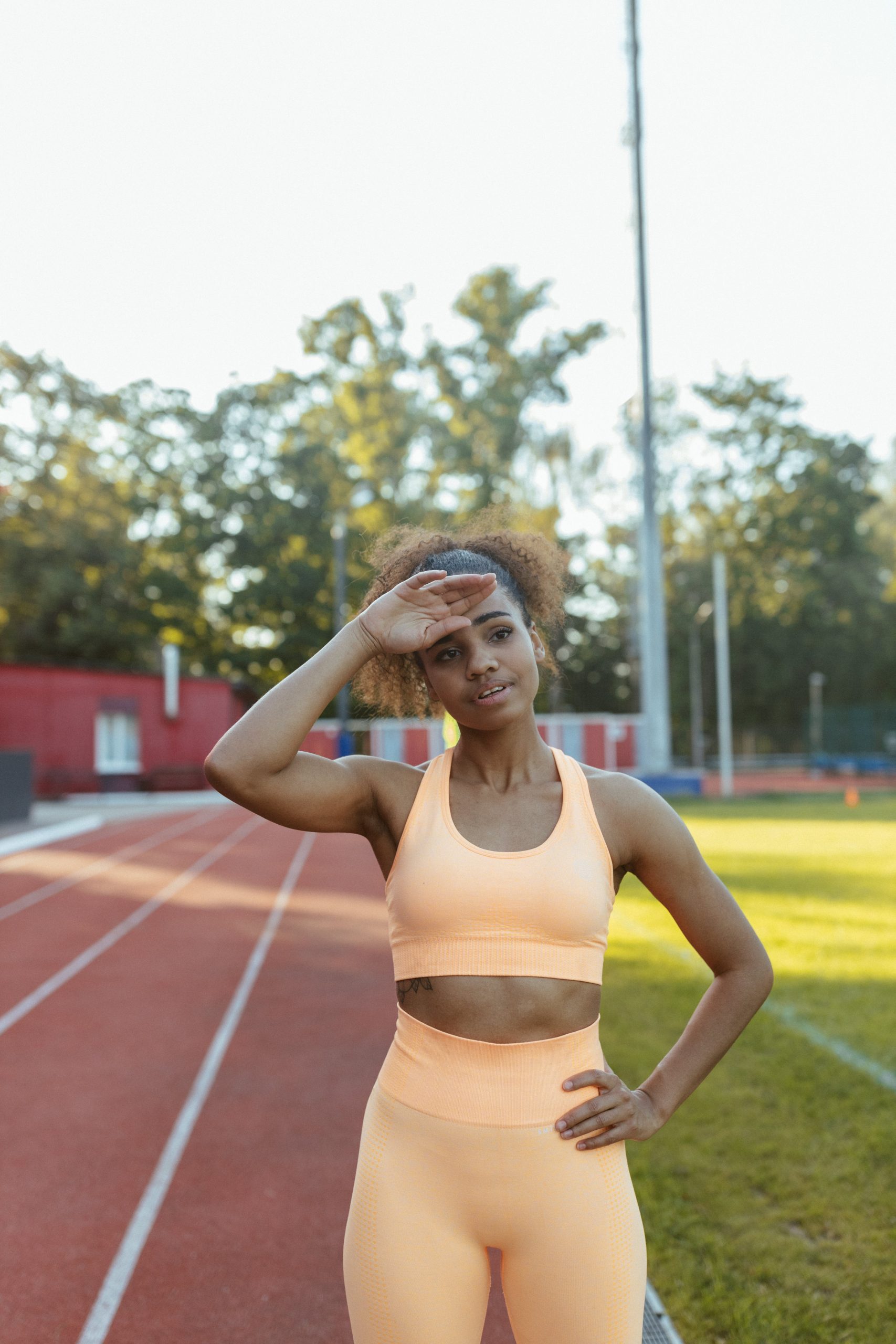 A female athlete in a peach sports outfit wiping sweat from her forehead on a sunny track field.