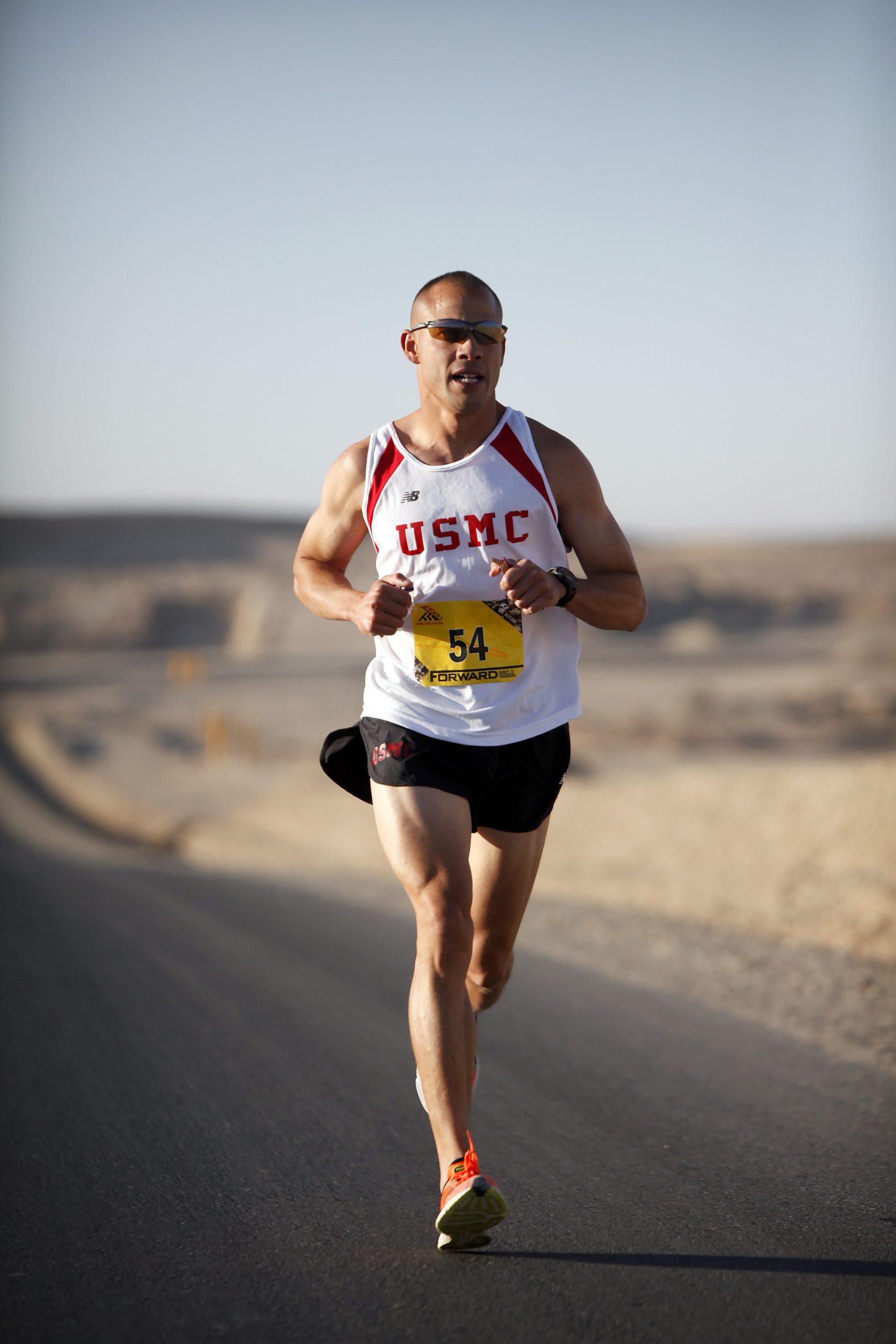 A runner with the letters "usmc" on his shirt and a race bib numbered 54 competes in a marathon on an asphalt road in a desert-like environment.
