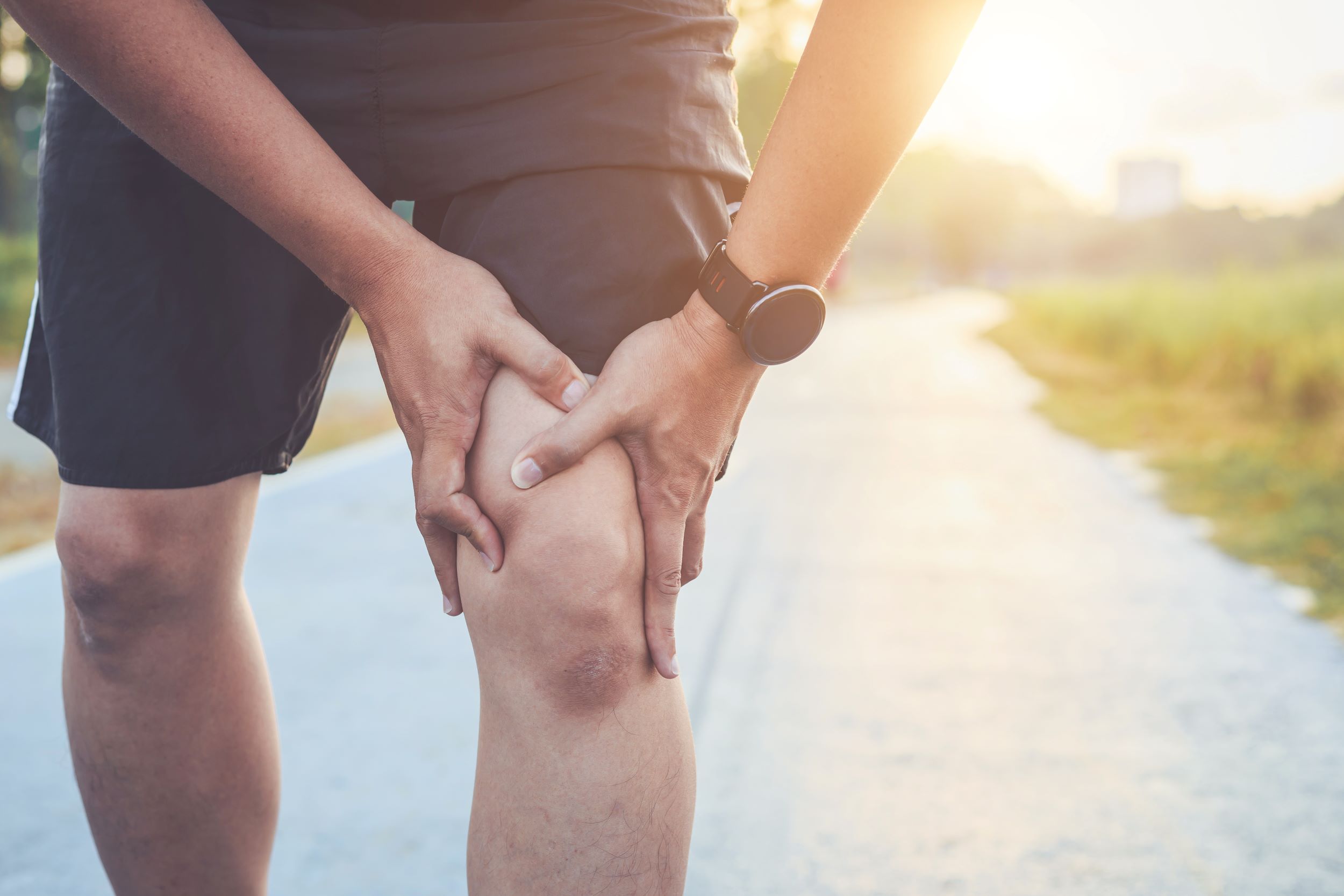 Person wearing a watch holding their knee with both hands in apparent discomfort while standing on an outdoor pathway in sunlight.