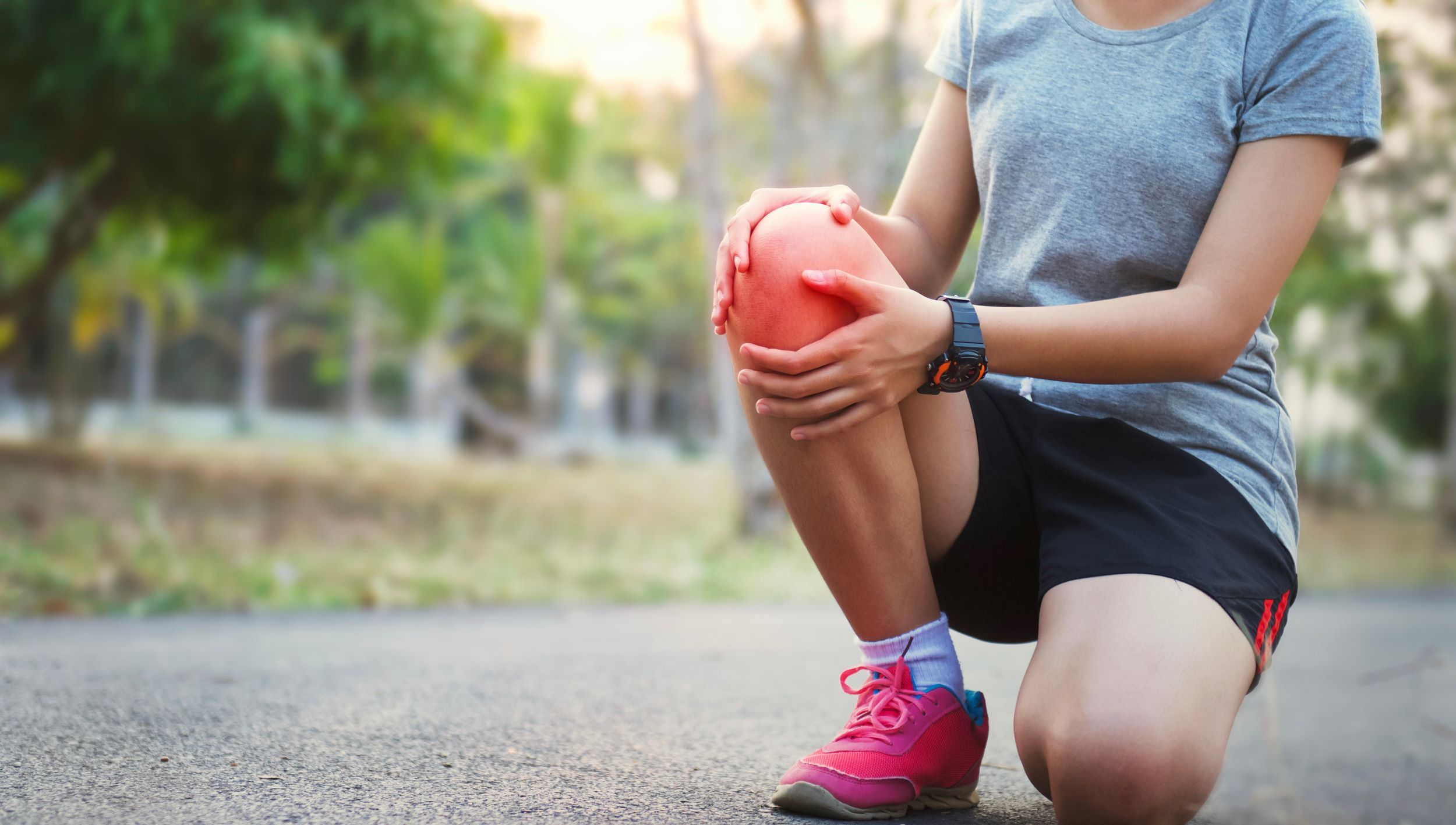 Person kneeling on a paved path holding their knee while wearing athletic clothing, a watch, and pink running shoes. Trees and greenery are visible in the background.