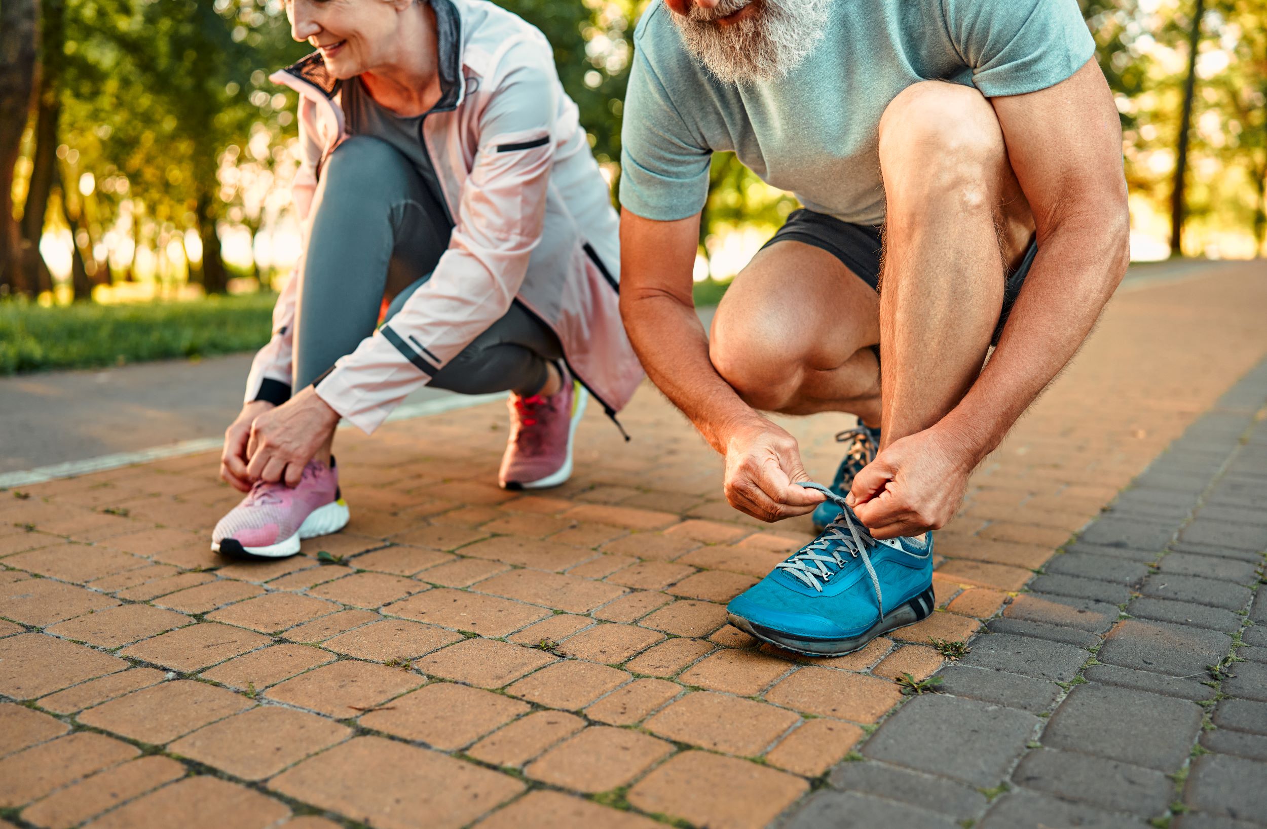 Two people in athletic wear are tying their shoes on a brick-paved path in a park. One is wearing a pink jacket and shoes, and the other is wearing a grey shirt and blue shoes.