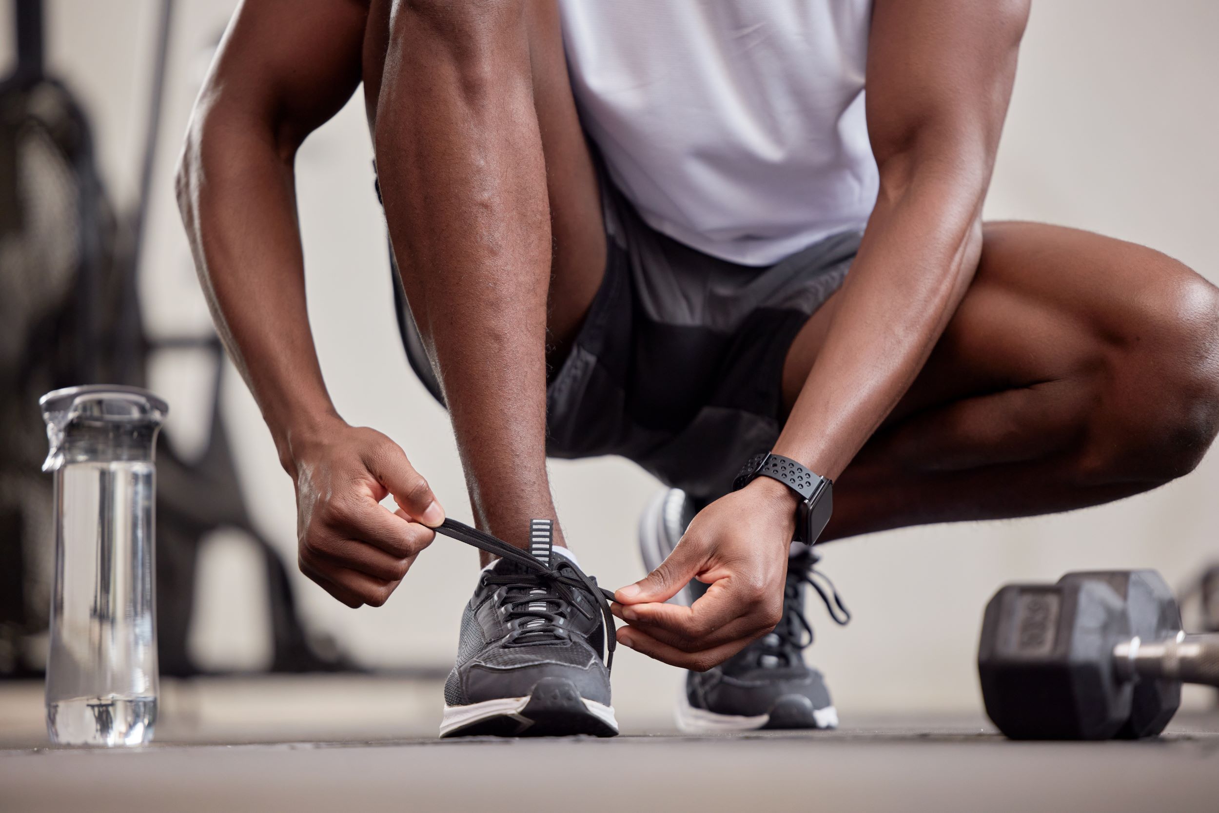 A person in athletic gear crouches to tie their sneaker, with a water bottle and a dumbbell next to them.