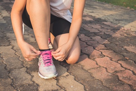 A person kneels to tie the pink laces on a gray sneaker while on a paved surface outdoors.