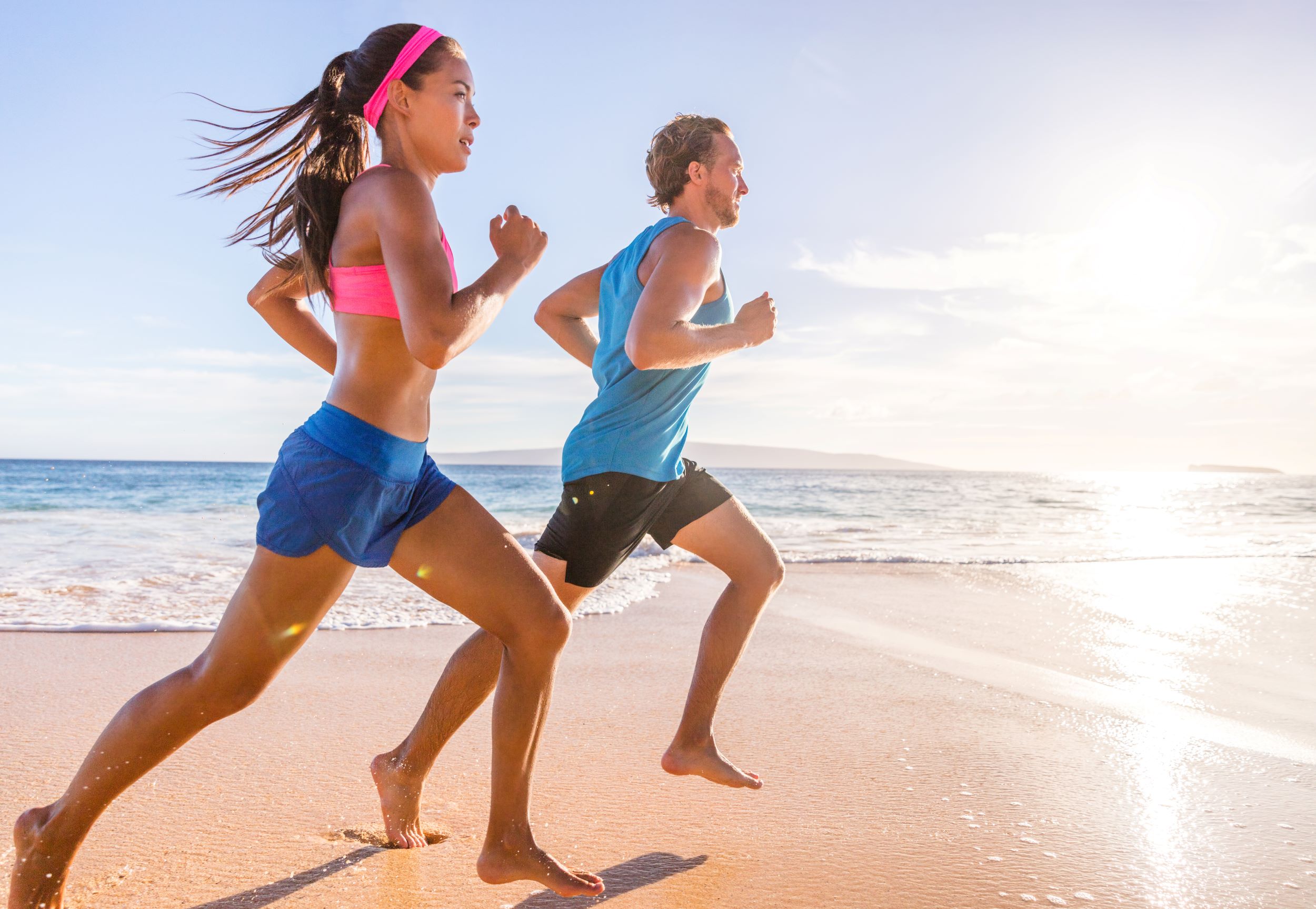 Two people are jogging barefoot on a sandy beach near the water's edge under a clear sky, with the sun shining brightly.