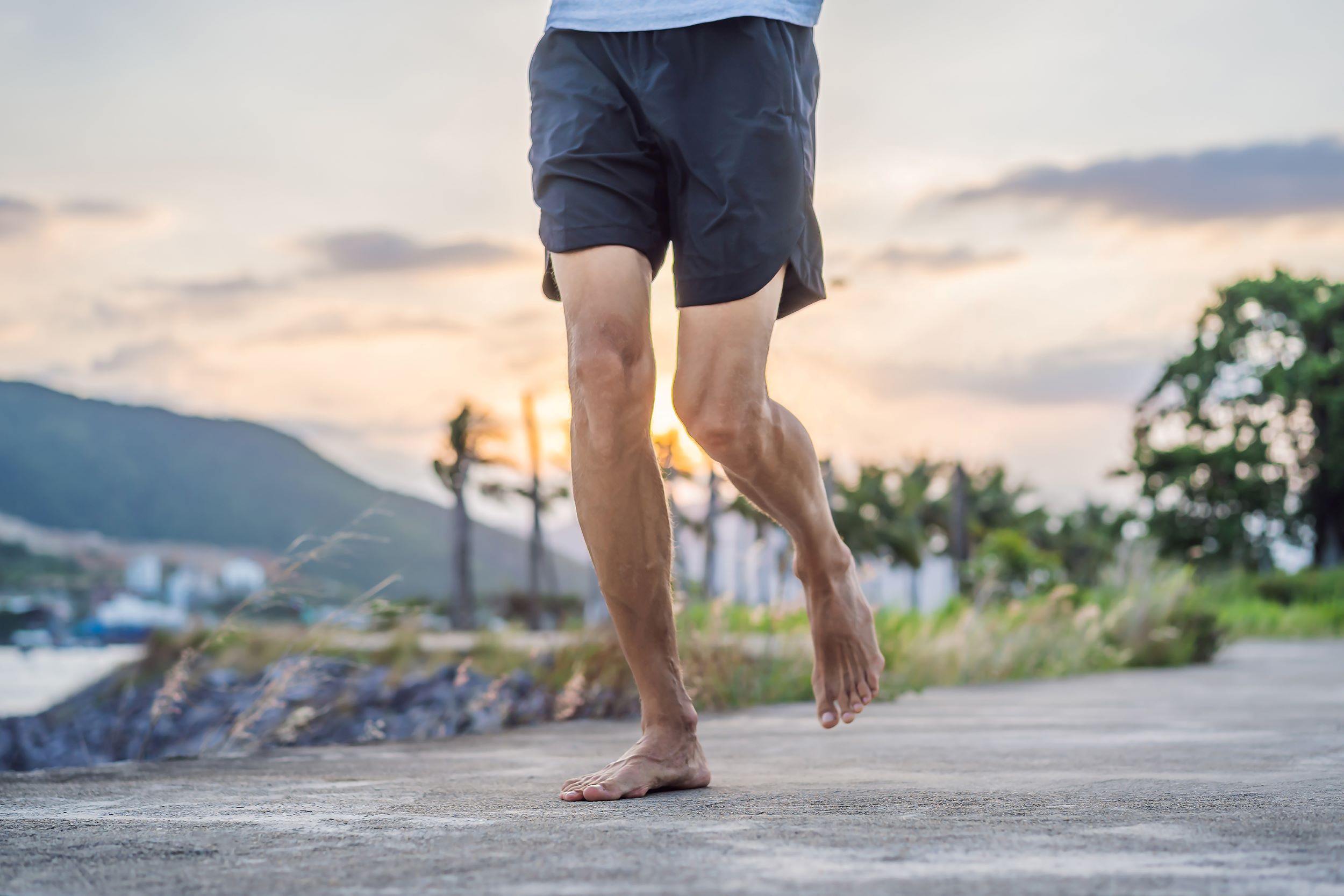 A person with bare feet running on an outdoor path with a scenic mountain and sunset background.