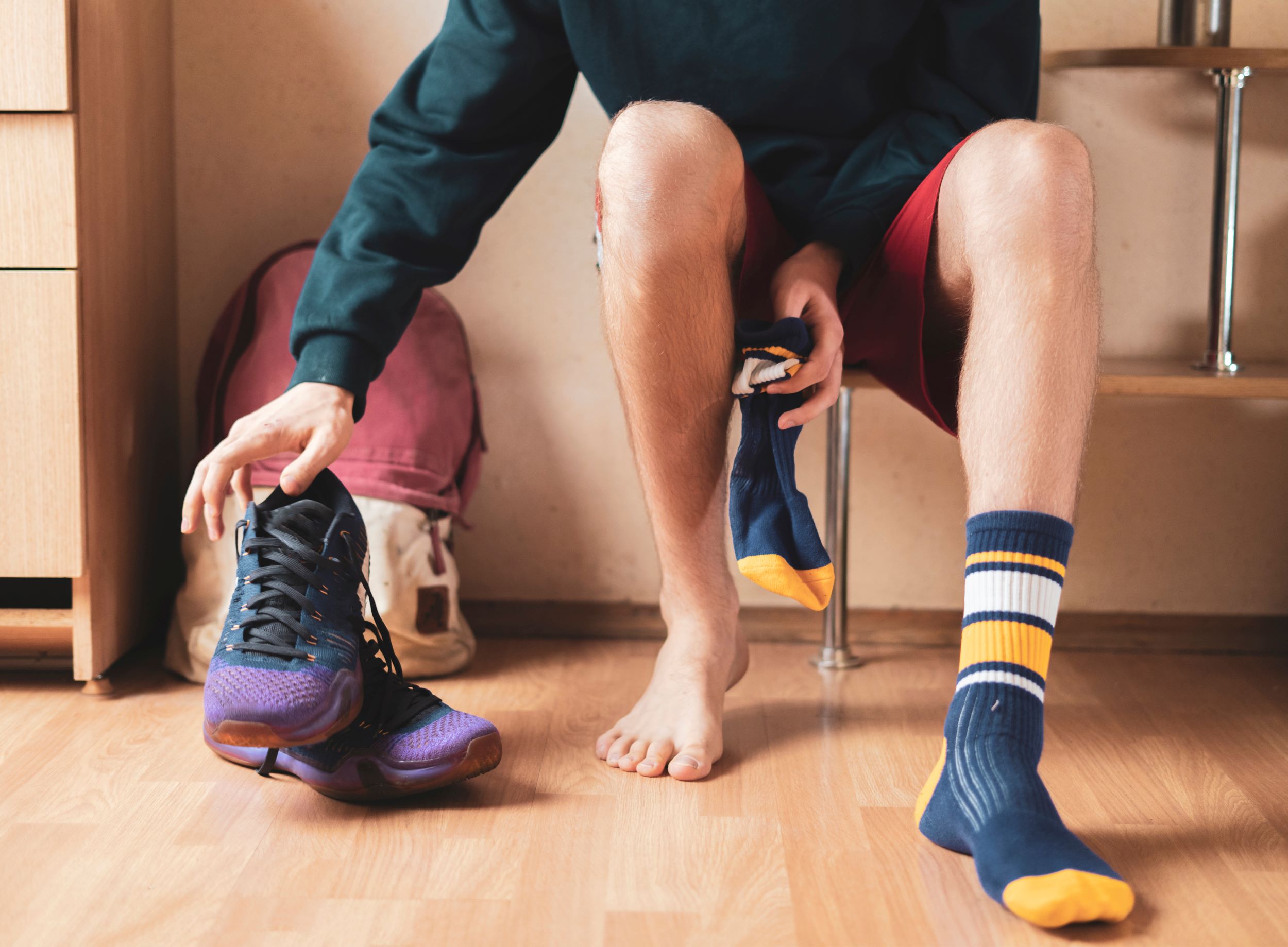 Person sitting on a chair, putting on a blue sock with yellow stripes, holding a purple athletic shoe in one hand, with a red backpack in the background.