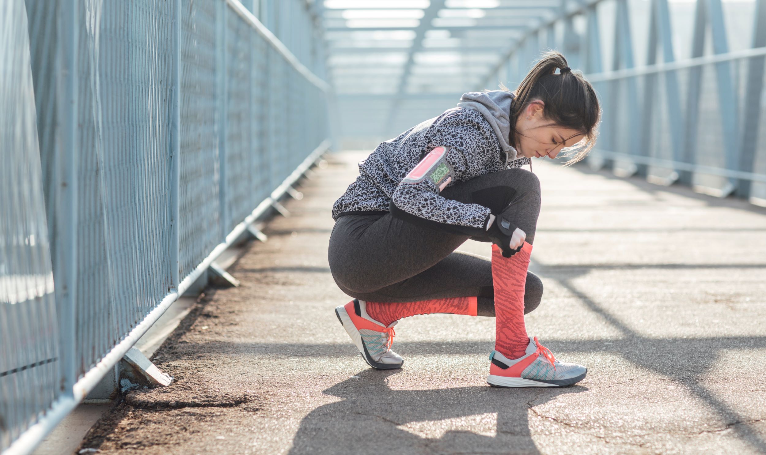 A woman in athletic wear ties her shoe while kneeling on a pedestrian bridge. She wears a gray hoodie, black leggings, and red sneakers. Sunlight casts shadows across the path.