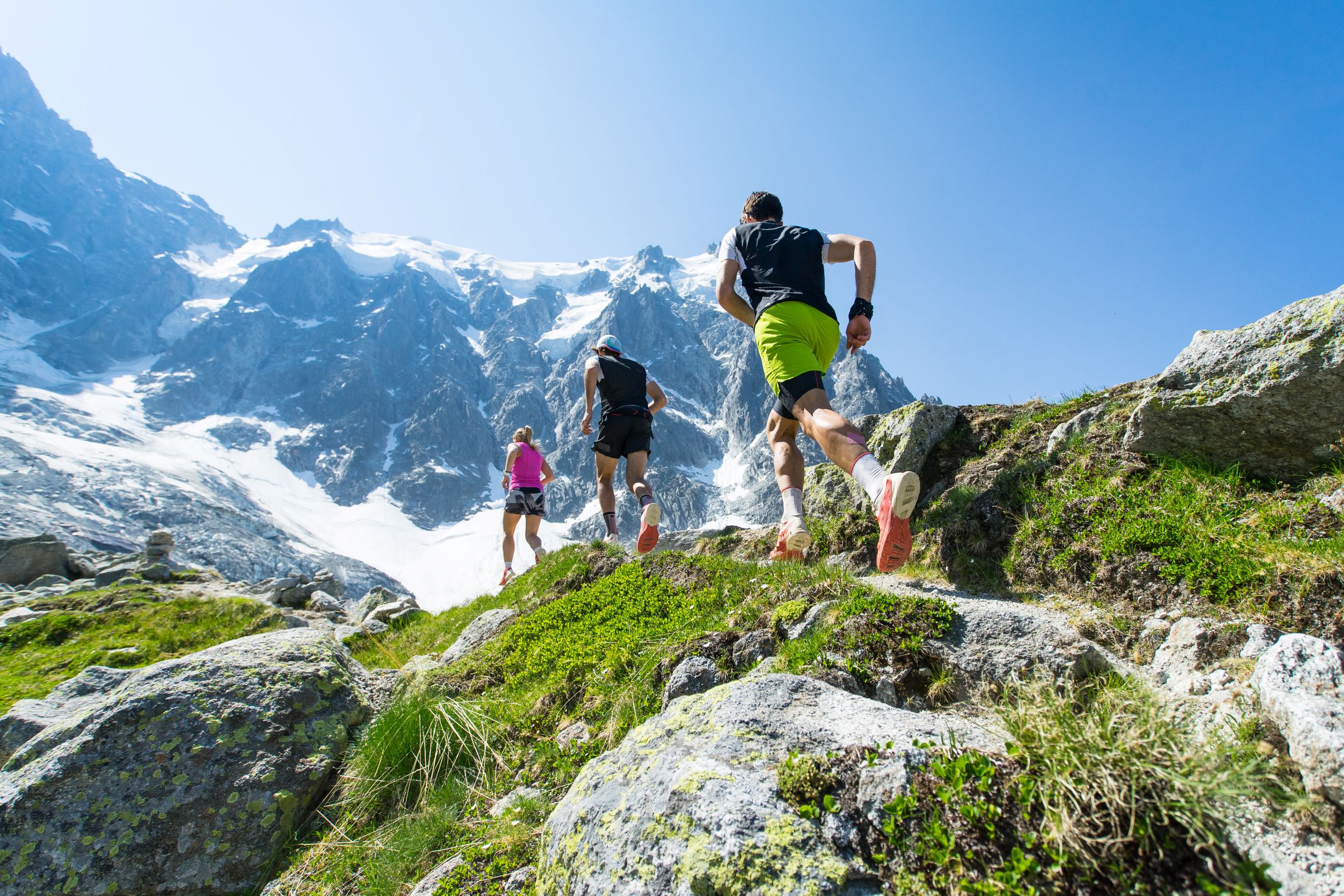 Three people trail running uphill on a rocky path with snow-capped mountains in the background on a clear, sunny day.